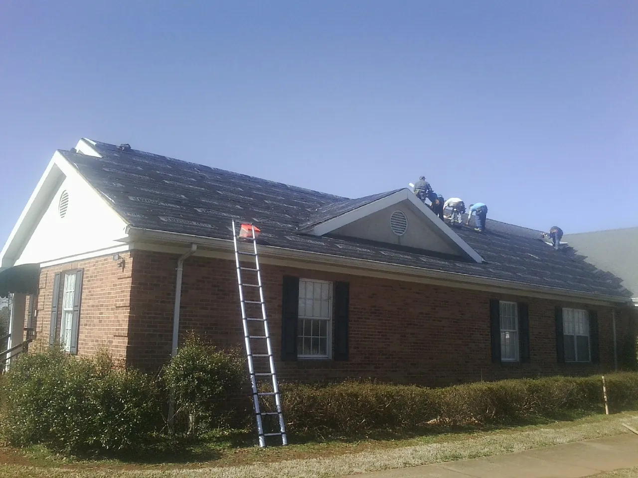 Crew tearing shingles off a brick ranch roof with a ladder set — Emergency Roof Repair work by our crew in Walnut Cove